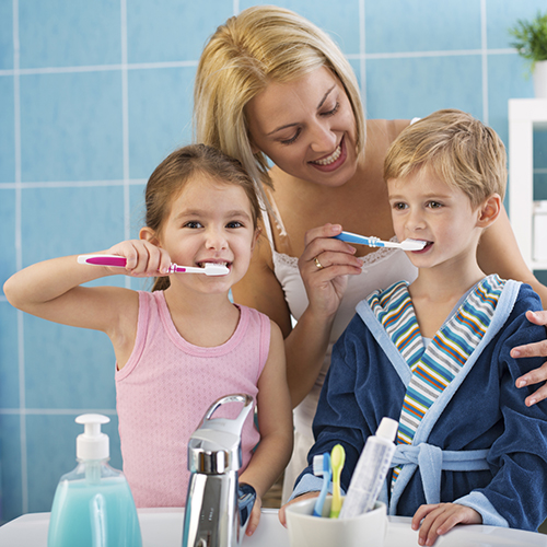 A woman in a bathroom brushing the little boy’s teeth and a little girl beside him holding a toothbrush and toothpaste
